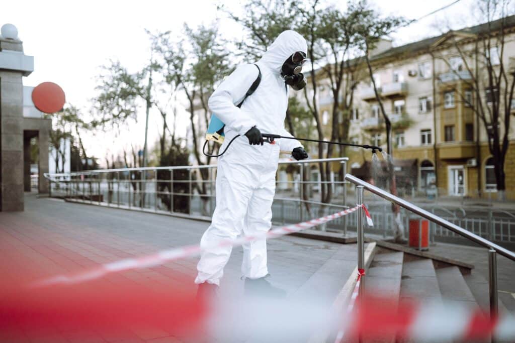 Man in protective suit and mask sprays disinfector onto the railing in the public place. Covid -19.